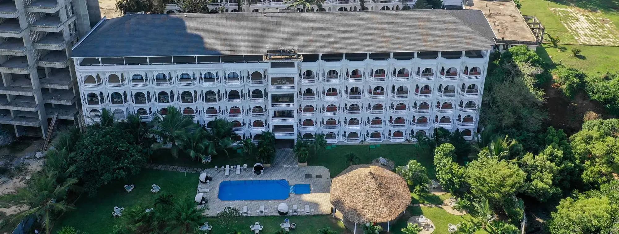 Aerial view of a white multi-storey hotel with arched balconies, a swimming pool and a thatched pavilion in a garden