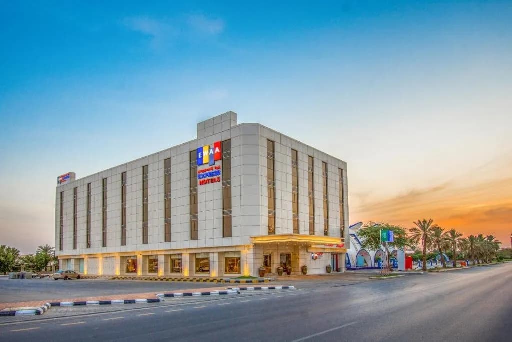 Exterior of a modern city hotel with a lit entrance at sunset, with palm trees beside the road